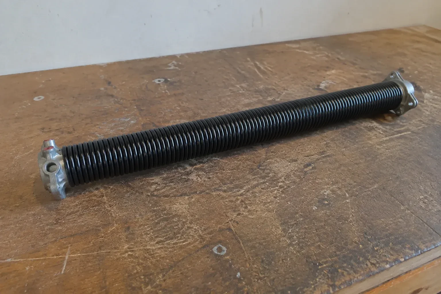 Detached black coil spring with metal end fittings on a workbench. — Torsion Spring Replacement, Cobourg